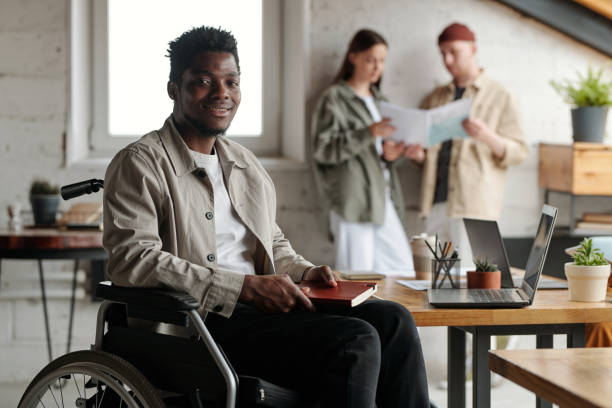 Happy young African American male broker with disability sitting in wheelchair in front of camera in loft coworking space and looking at you