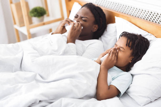 Respiratory Disease. Black mom and daughter sneezing, blowing noses on paper napkins in bed