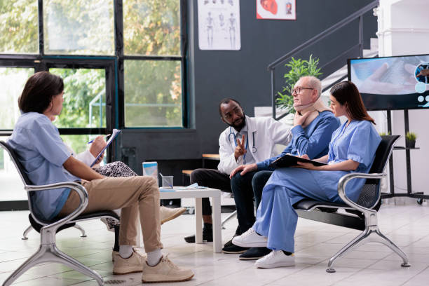 Diverse medical team discussing with injured elderly patient to remove neck collar during consultation in hospital waiting room. Asian assistant holding clipboard writing health care treatment