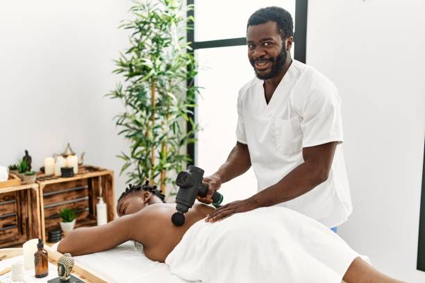 African american woman reciving back massage with gun percusion at beauty center.