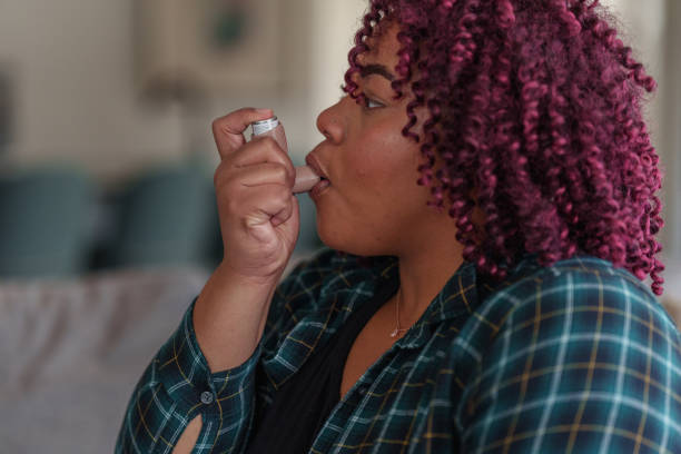 A multiracial woman sits on the sofa at home and uses an inhaler to treat asthma and allergies.