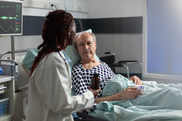 African american therapist in hospital room discussing treatment holding pills bottle reassuring recovery,sick senior man laying in bed breathing through oxygen mask,