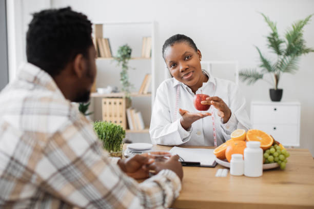 Female nutritionist holding apple with measuring tape while sitting at office desk in front of adult patient. Attractive multiracial woman in white coat promoting healthy eating with slimming foods.