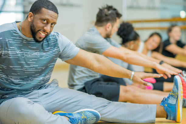 A multi-ethnic group of adult men and women are indoors in a gym. They are wearing casual exercise clothing and running shoes. They are doing stretches by touching their toes. A man of African descent is smiling at the camera.