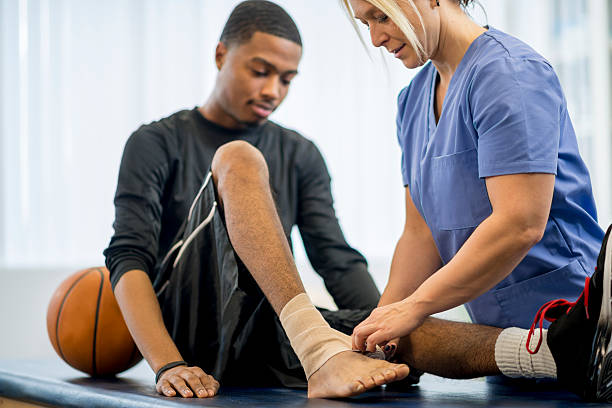 A male teenage basketball player is at the doctors office to have some work done on his injury. A nurse is wrapping his bandage around the ankle.