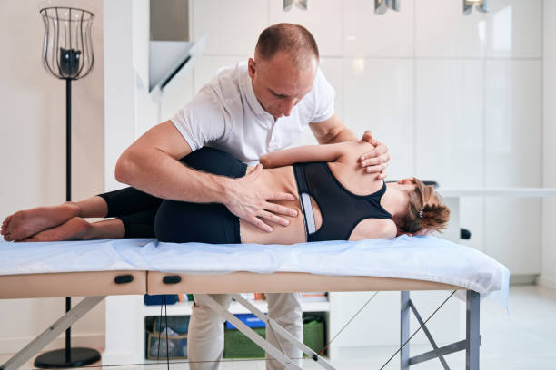 Waist up front view portrait of strong man physiotherapist in white shirt stretching woman back on medical couch in wellness center