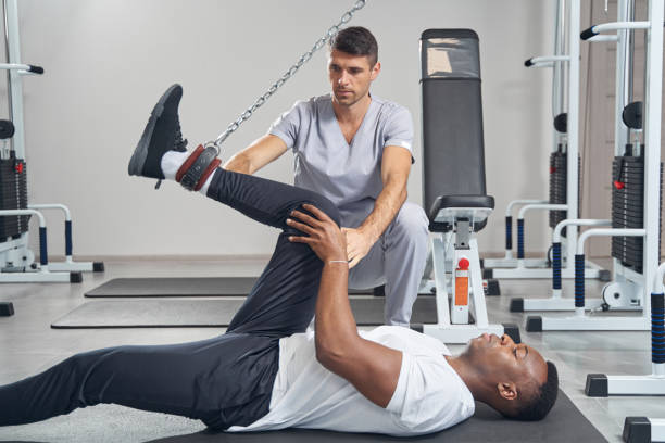 Male patient doing knee flexion on gym equipment assisted by qualified physical therapist