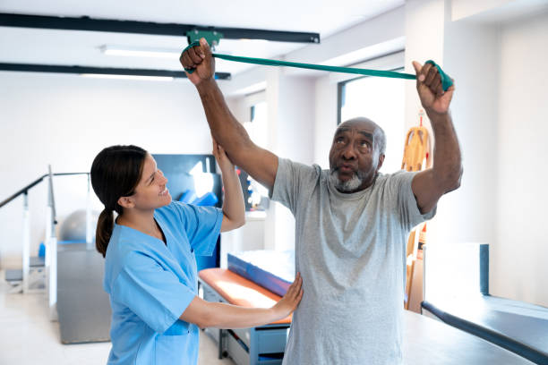 Black senior man working out his arms and shoulders using a resistance band and cheerful therapist standing next to him correcting his posture - healthcare concepts