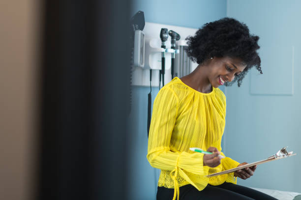 A black woman is at a routine medical appointment. The patient is sitting on a medical examination table in a clinic. She is filling out health history and medical insurance paperwork on a clipboard.