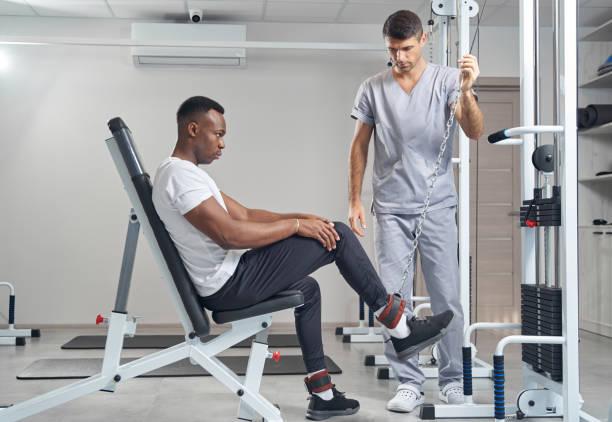 Serious concentrated young man performing seated leg raise assisted by experienced kinesiologist