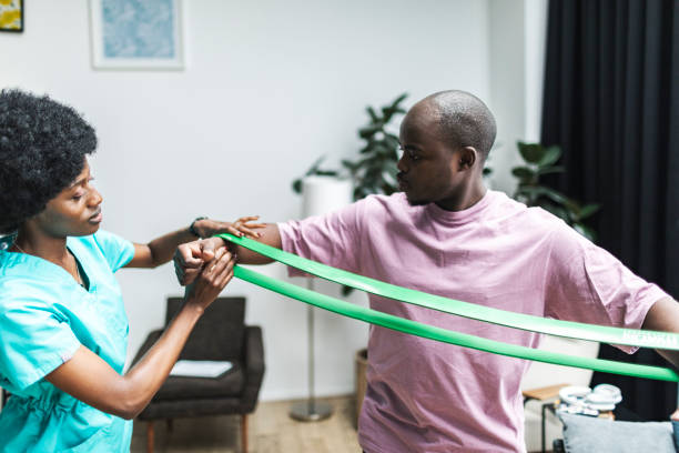 Young black physiotherapist working with patient at home doing exercises with exercise band, helping to recover after surgery