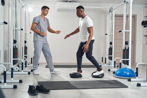 Man with ankle weights working out on two soft balance discs supervised by kinesiologist