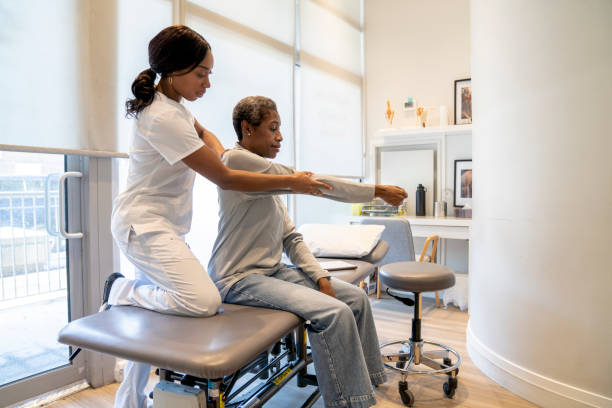 A senior woman of African decent, sits over the edge of the treatment table as her female Chiropractor carefully adjusts her back.  The Chiropractor is dressed professionally in white scrubs and is manually placing her patients arm in the proper position for the adjustment as she verbally walks her through the process.