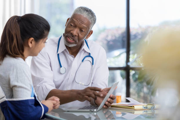 A serious senior male medical doctor sits at a table in his office with an unrecognizable patient.  He points at his computer screen as he explains her symptoms.  She wears an arm sling.