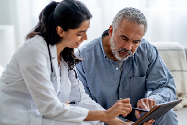 A female doctor meets with her patient in the comfort of his own home during a medical check up.  She is dressed professionally and reviewing recent test results with the gentleman on a tablet.