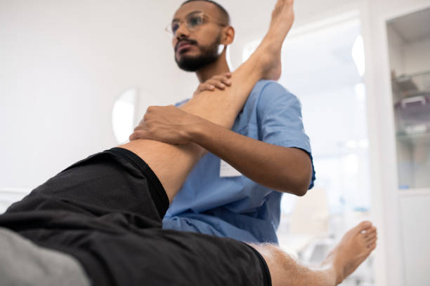 Qualified medical professional massaging sick knee of patient in hospital while holding his leg on shoulder