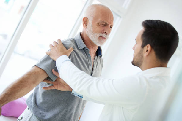 Closeup side view of an early 60's senior gentleman having some shoulder pain. He's at doctor's office having medical examination by a male doctor. The doctor is touching the sensitive area and trying to determine the cause of pain.